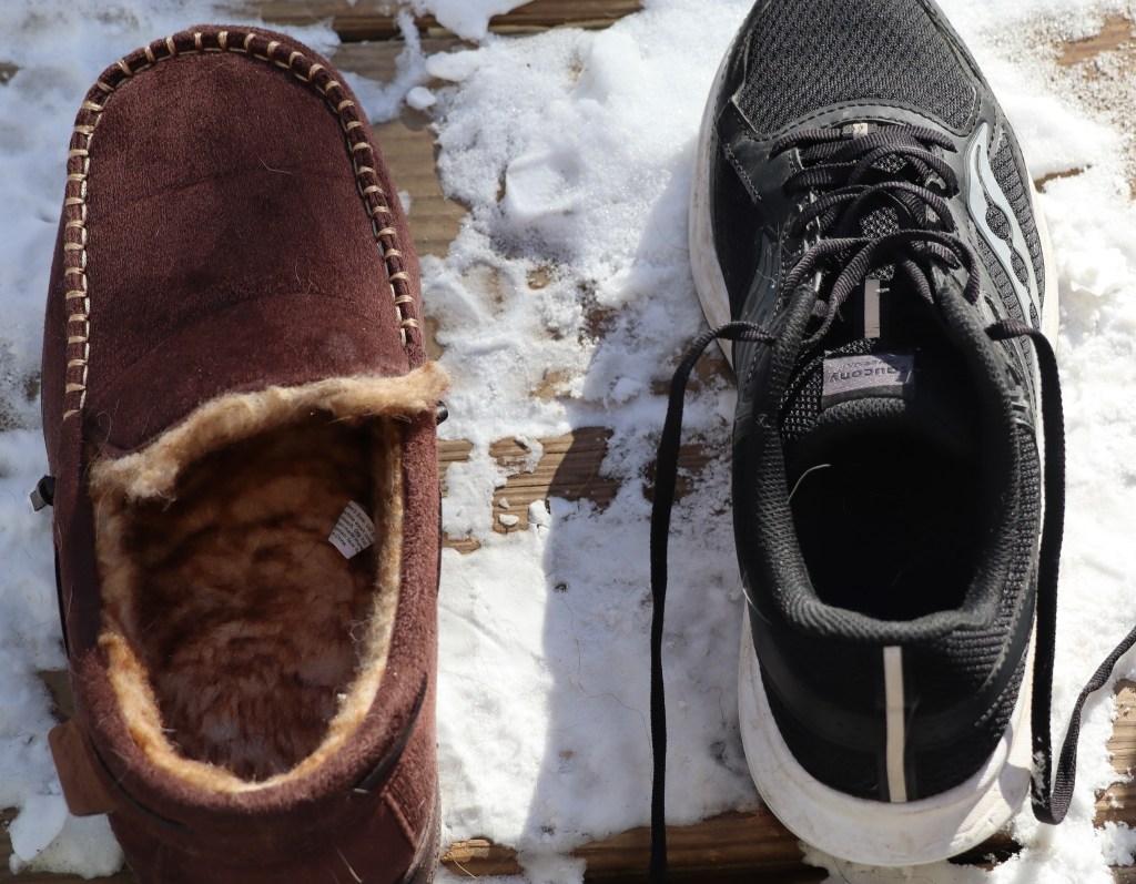 A comparison of a cozy brown slipper and a black running shoe placed on a snowy wooden surface.